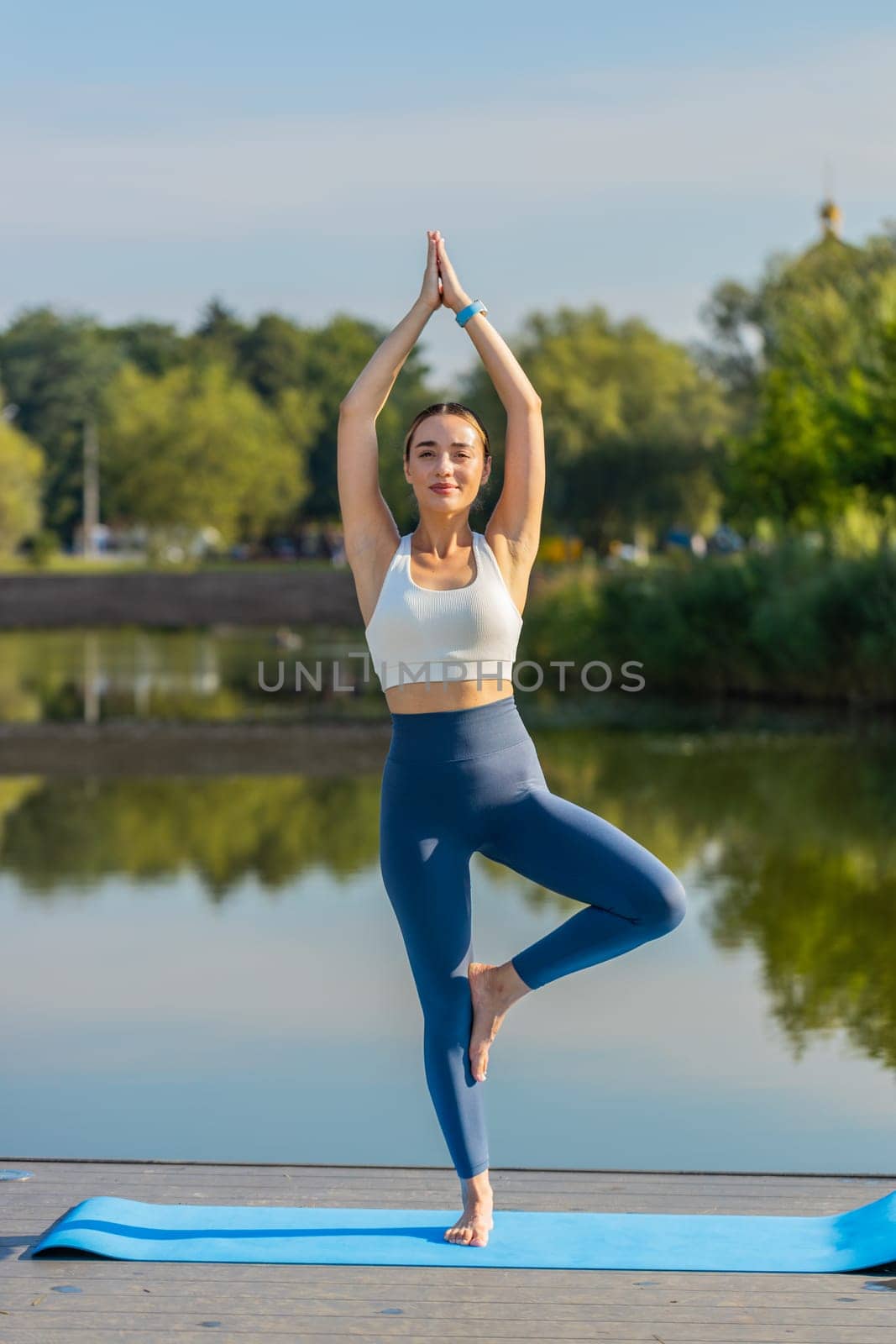 Caucasian slim flexibility calm sporty woman balance on one leg on mat near pond in park summer. Fit fitness young girl training wellbeing workout yoga female standing in Tree Pose. Healthy lifestyle.