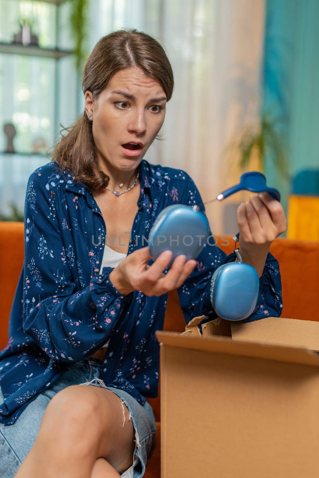 Unhappy young Caucasian woman unpacking carton box, feeling dissatisfied with getting damaged wireless headphones order, writing negative feedback in mobile application, internet shopping concept.