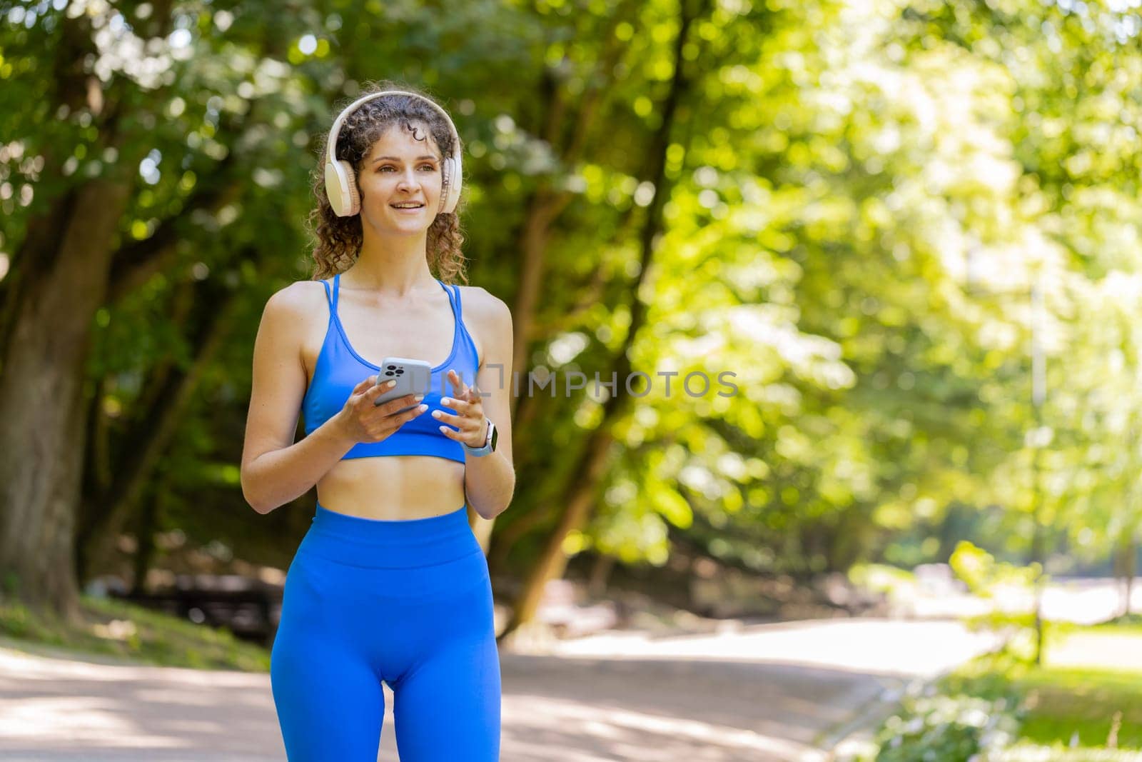 Athletic fitness adult girl sport runner woman walking, wearing headphones listening music from smartphone and smiling. Healthy sportswoman workout cardio outdoors in morning park on sunny summer day.