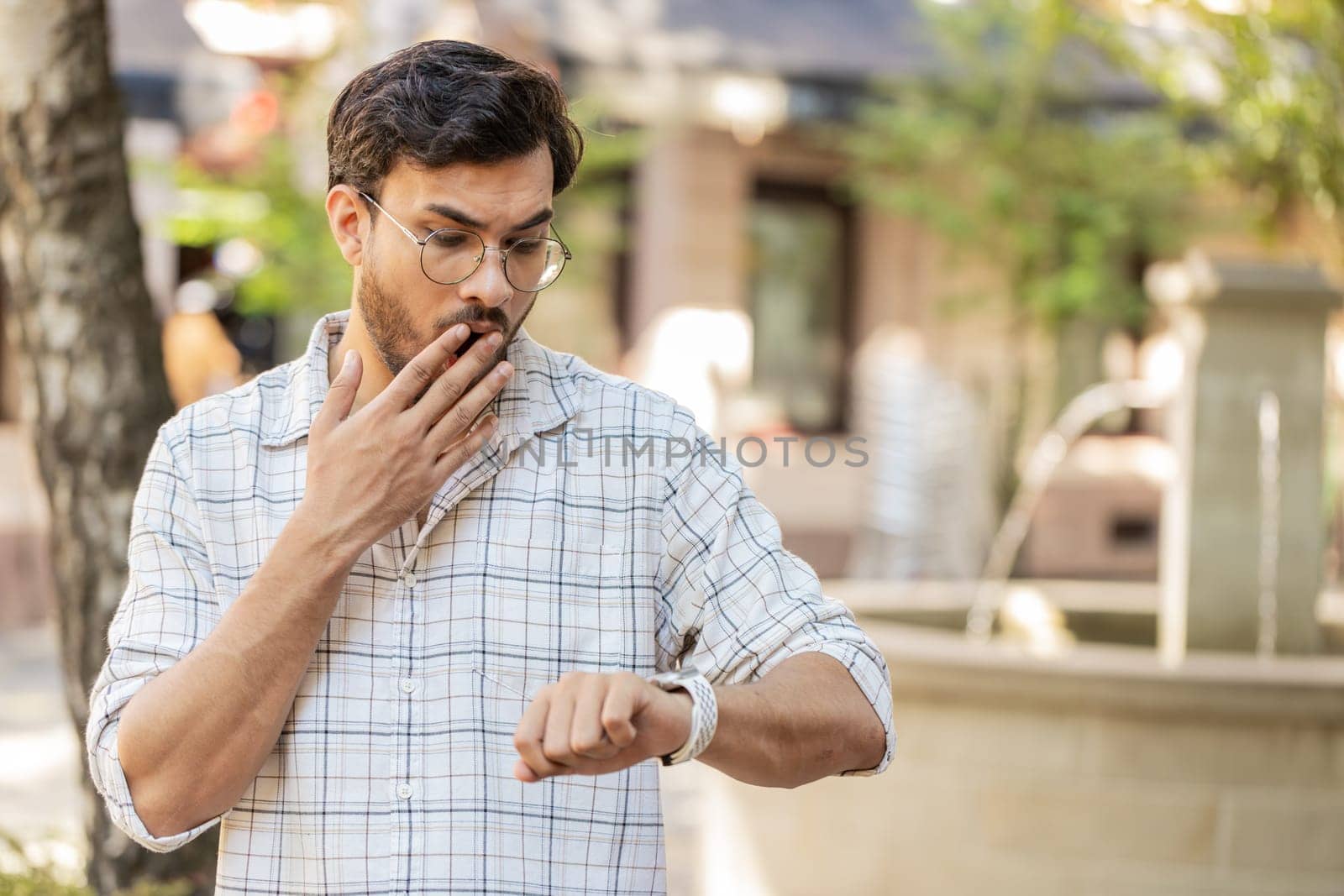 Scared man worrying to be punctual, with anxiety checking time on watch, running late to work or transport, being in delay, deadline outdoors. Indian guy tourist walking in city street. Town lifestyle