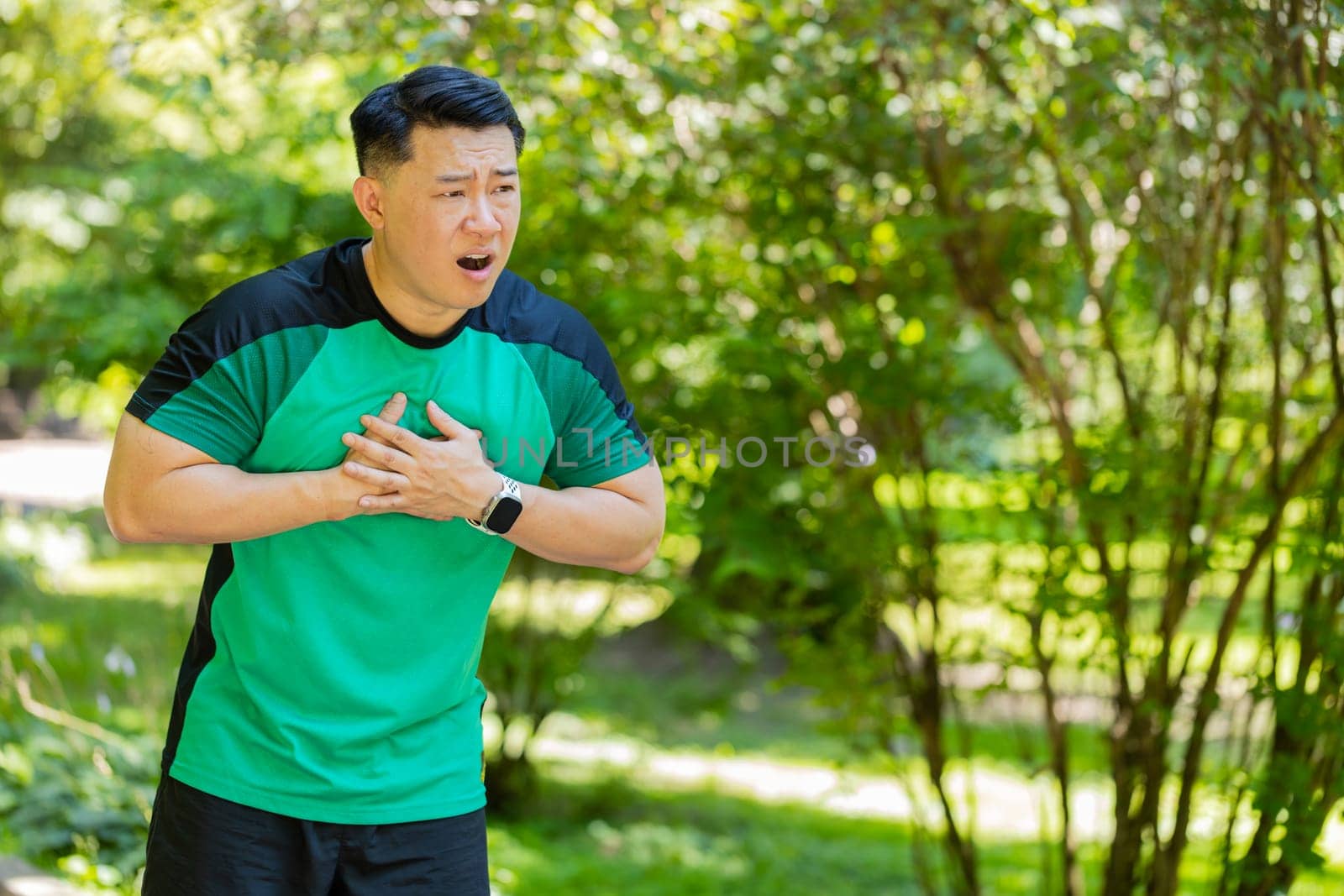 Young adult Asian runner having heart attack or chest pain after running workout in park on sunny summer day. Tired Japanese athlete suffers from tension in the heart. Health care and sports concept.