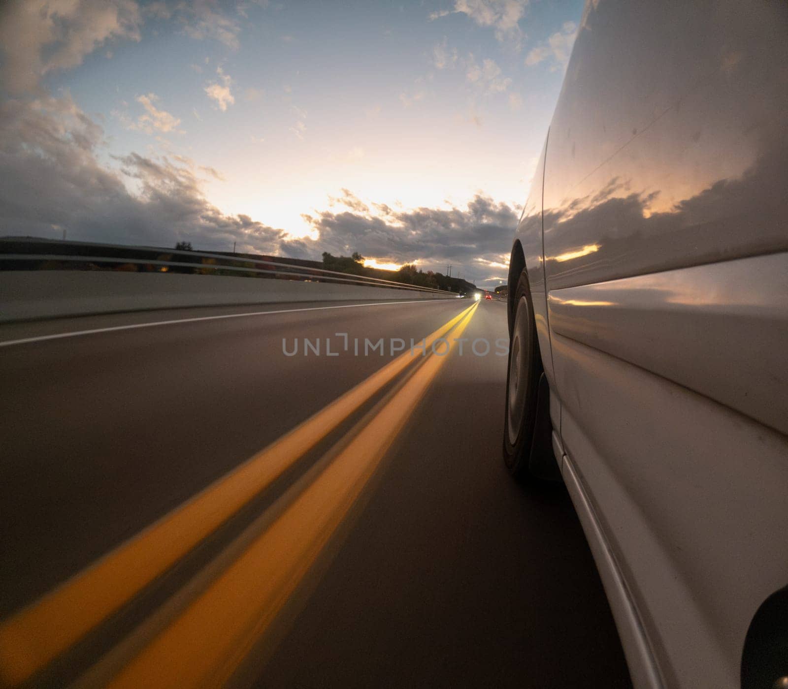 A car is driving down a road with a sunset in the background. The car is white and has its headlights on