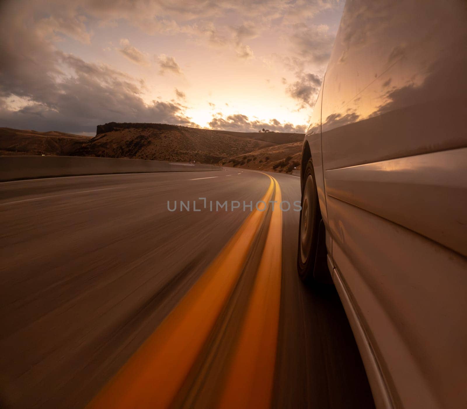 A car is driving down a road with a sunset in the background. The road is lined with yellow lines, and the car is moving quickly