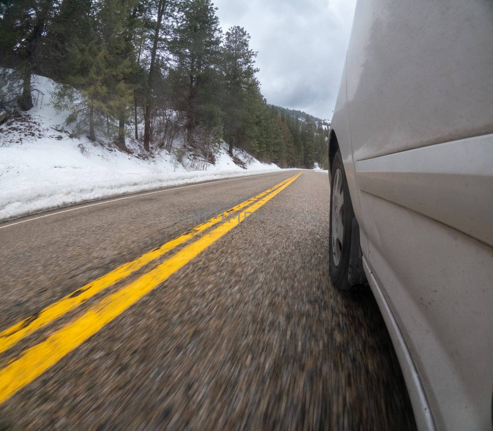 A car is driving down a snowy road with yellow lines. The car is white and has a tire that is visible. The scene is blurry, giving the impression of motion