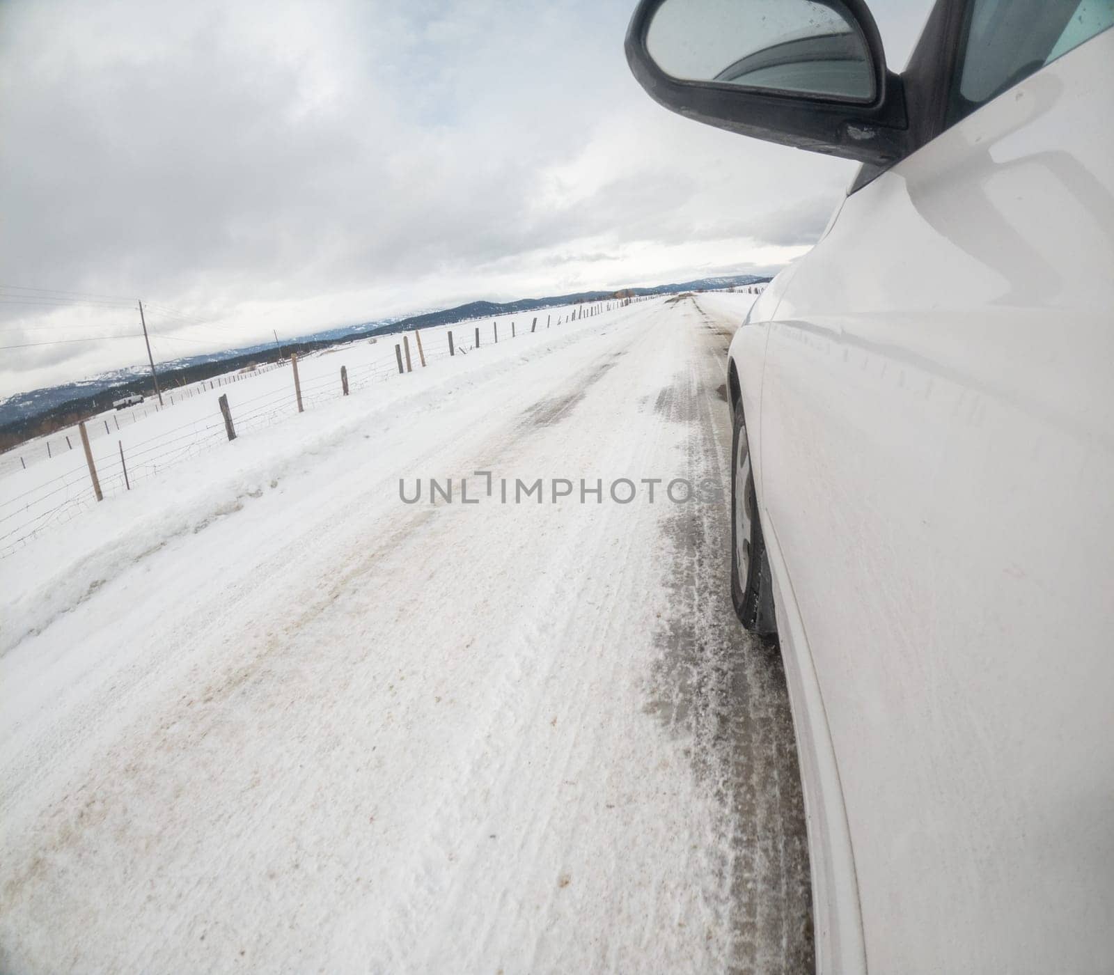 A car is driving down a snowy road with a fence in the background. The car is in the driver's mirror and the road is covered in snow