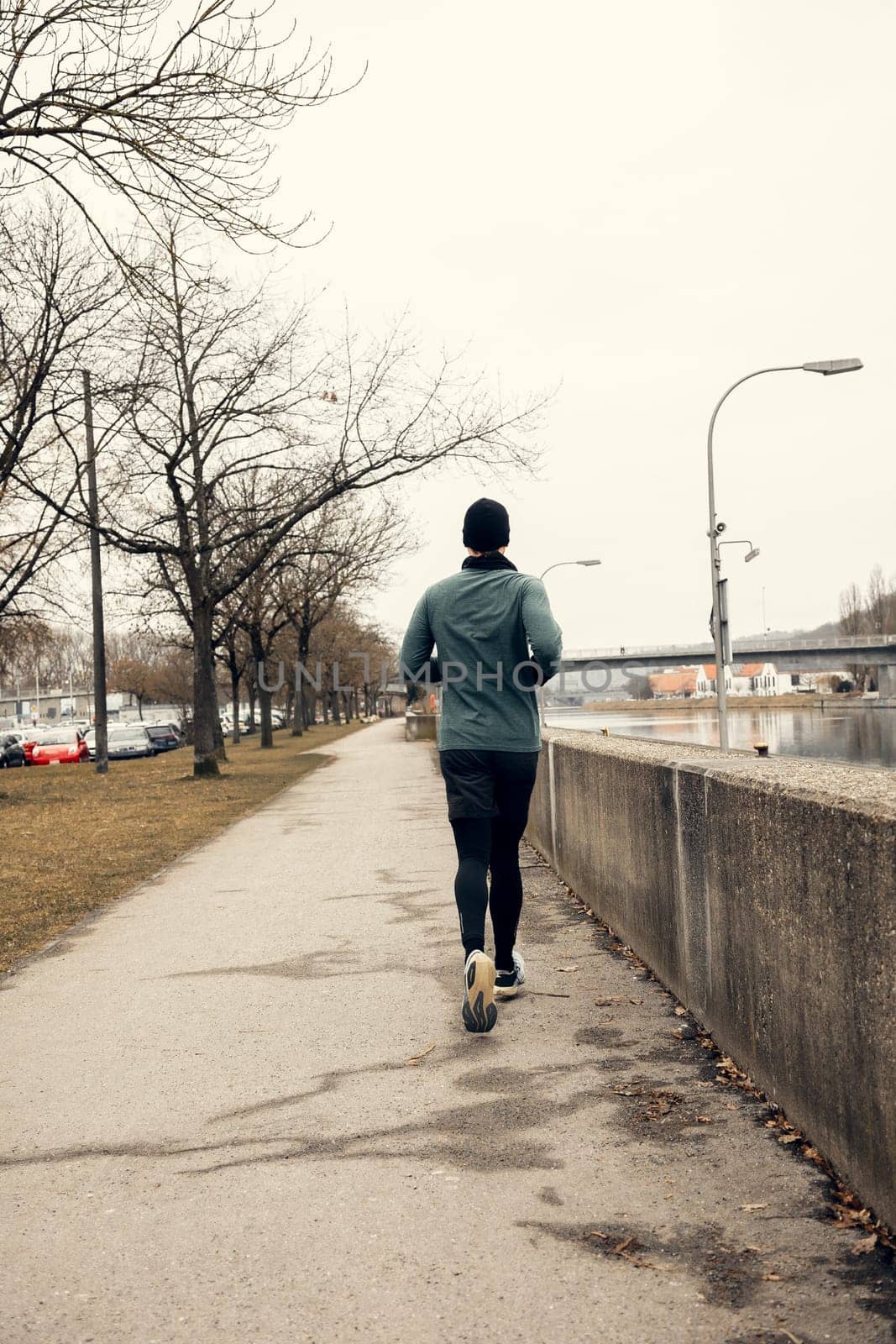 A man in sportswear jogging along a river on a cloudy day. The empty pathway, bare trees, and cityscape create a serene and urban fitness atmosphere