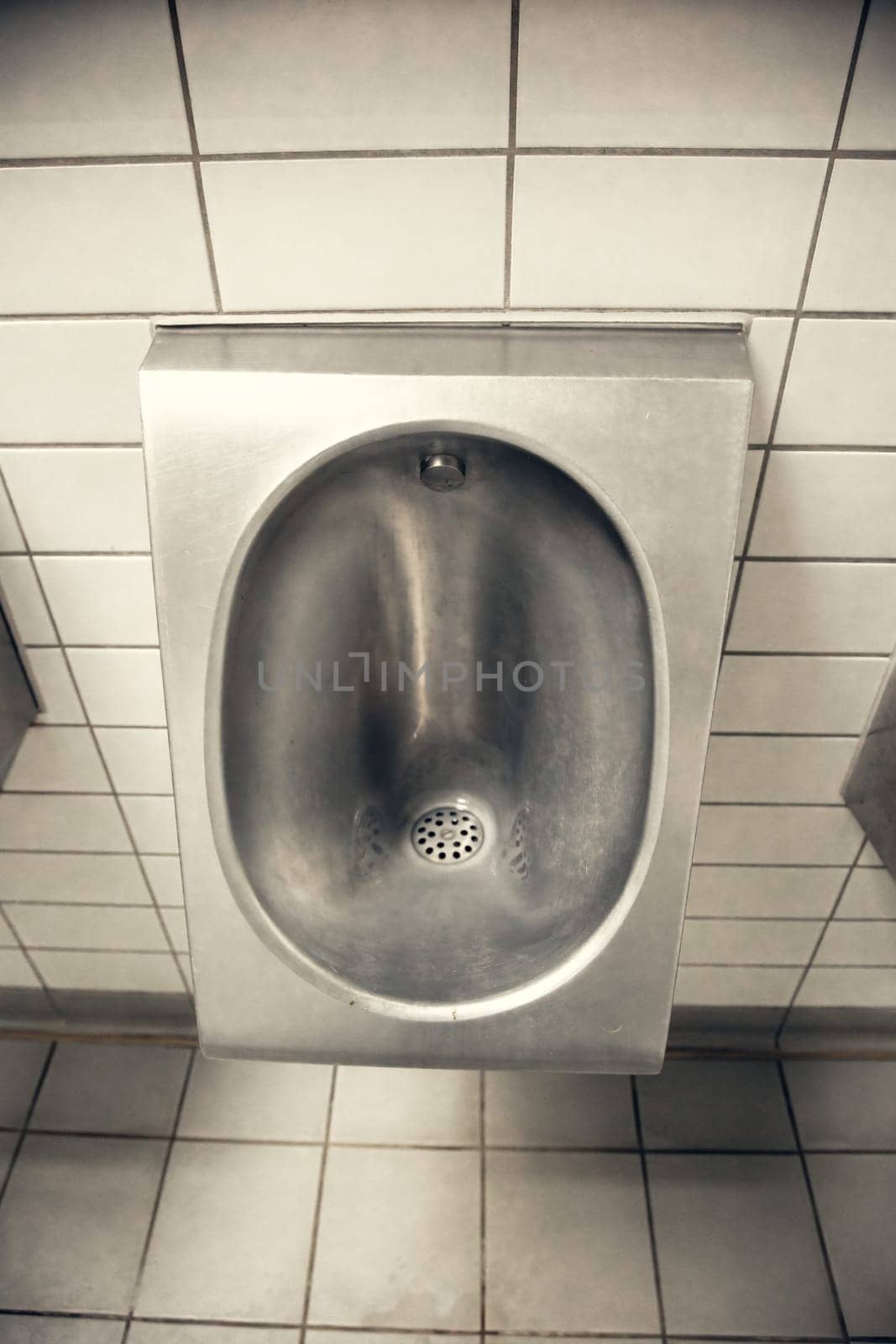 A stainless steel urinal installed in a public restroom with white tiled walls. Minimalist and industrial style, emphasizing hygiene, cleanliness, and functional design