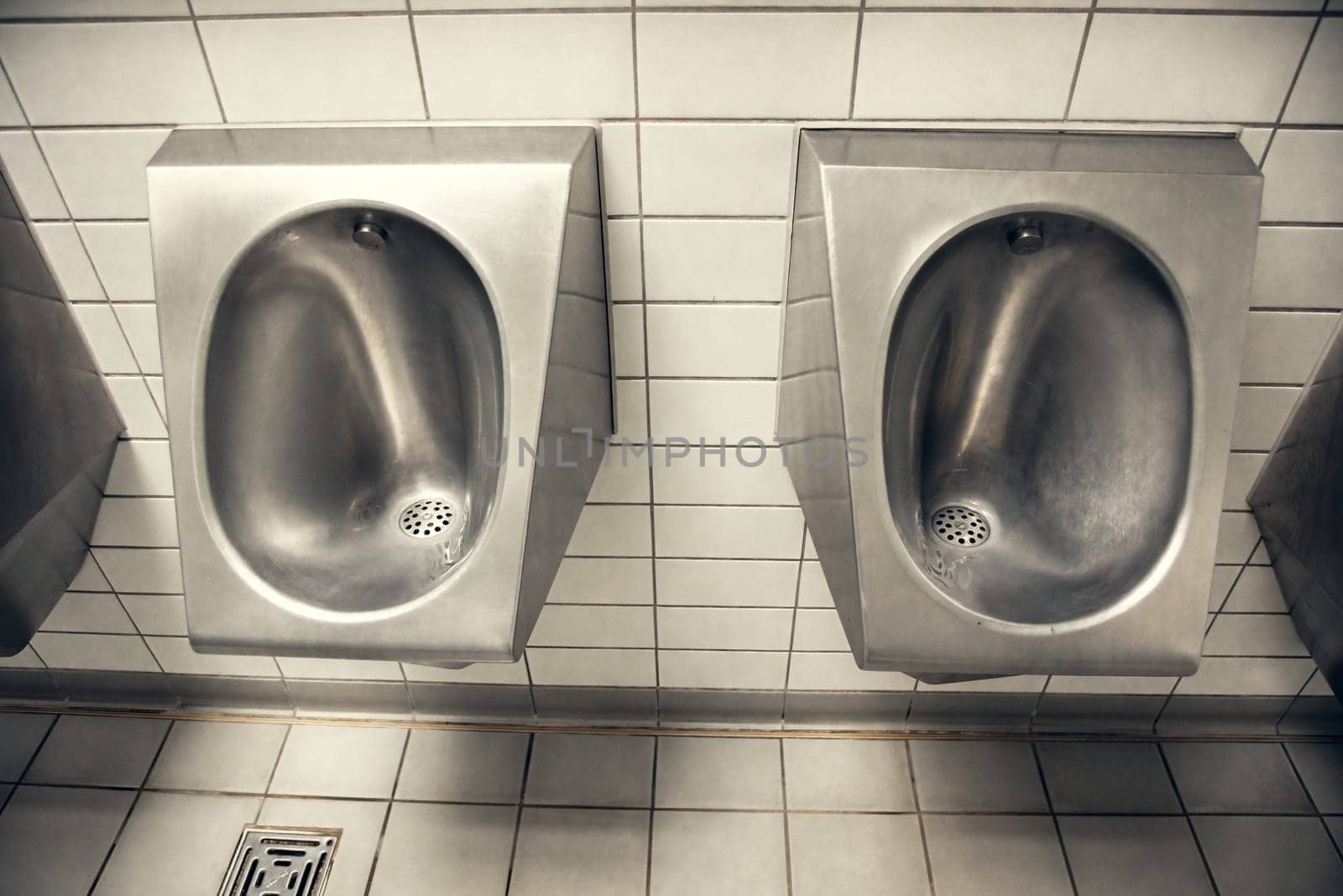 Stainless steel urinals in a public restroom with white tiled walls. The industrial and functional design is perfect for hygiene-related themes, public facilities, and restroom interiors