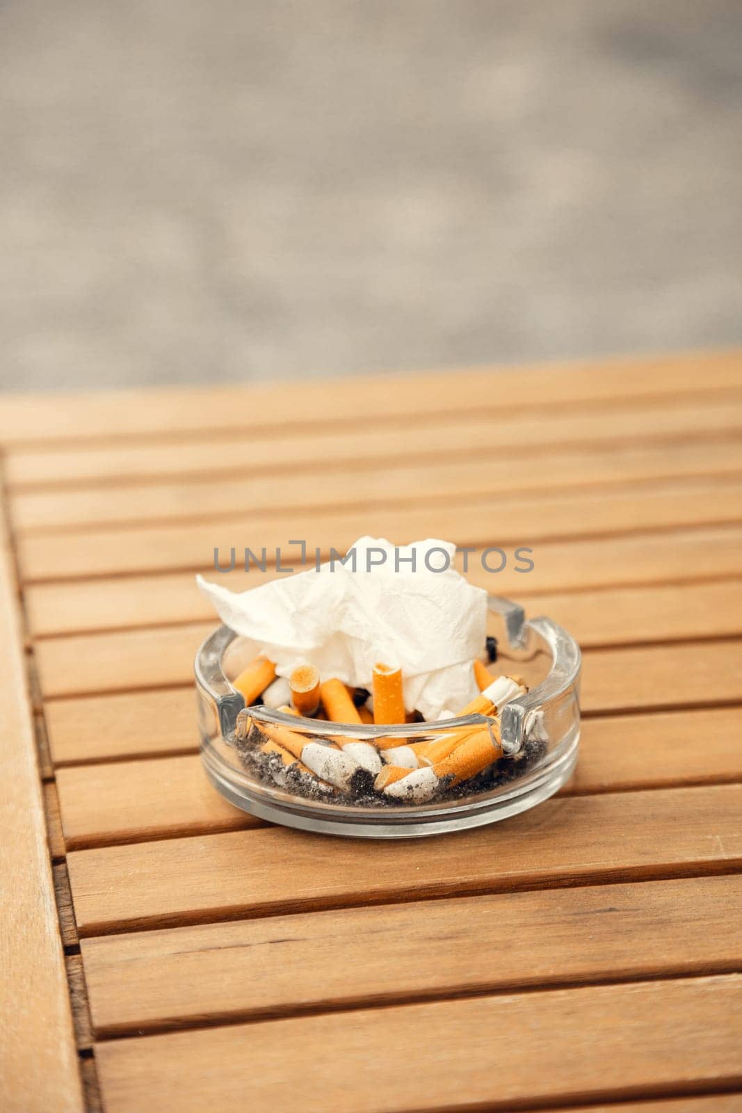 A glass ashtray filled with cigarette butts and tissue on a wooden cafe table. A detailed close-up highlighting smoking culture, environmental concerns, and public space hygiene