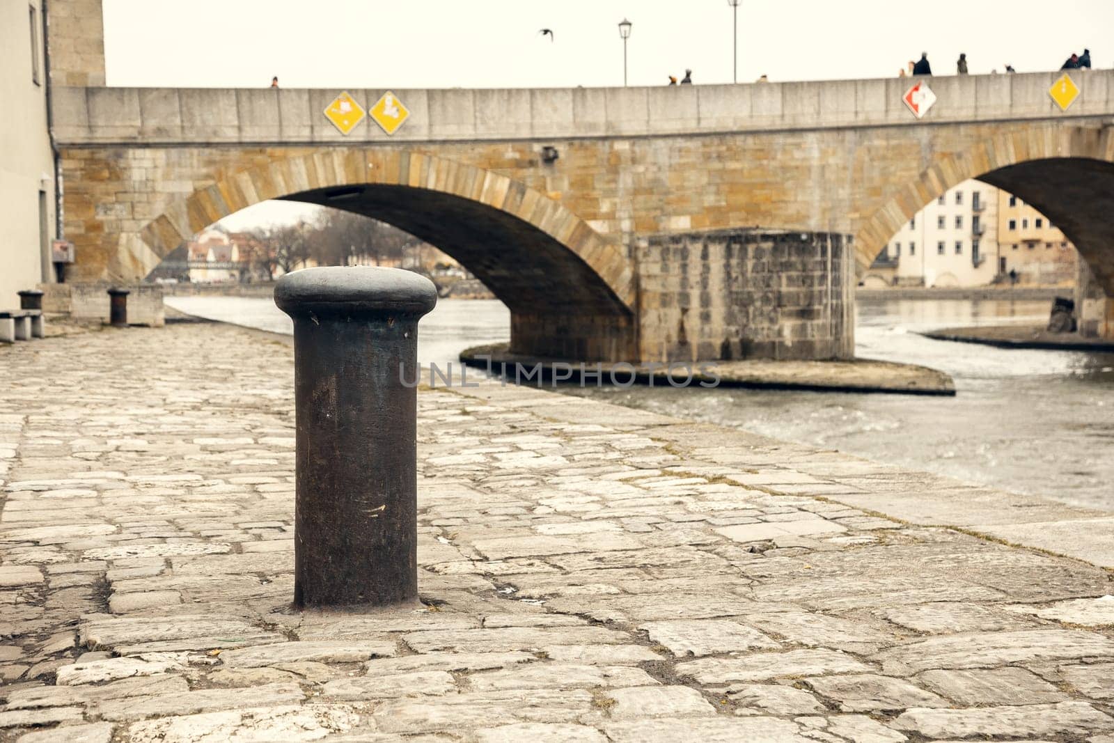 A rustic mooring bollard on an old cobblestone riverbank, with a historic stone bridge spanning the river in the background. A perfect image for travel, history, and urban heritage themes.