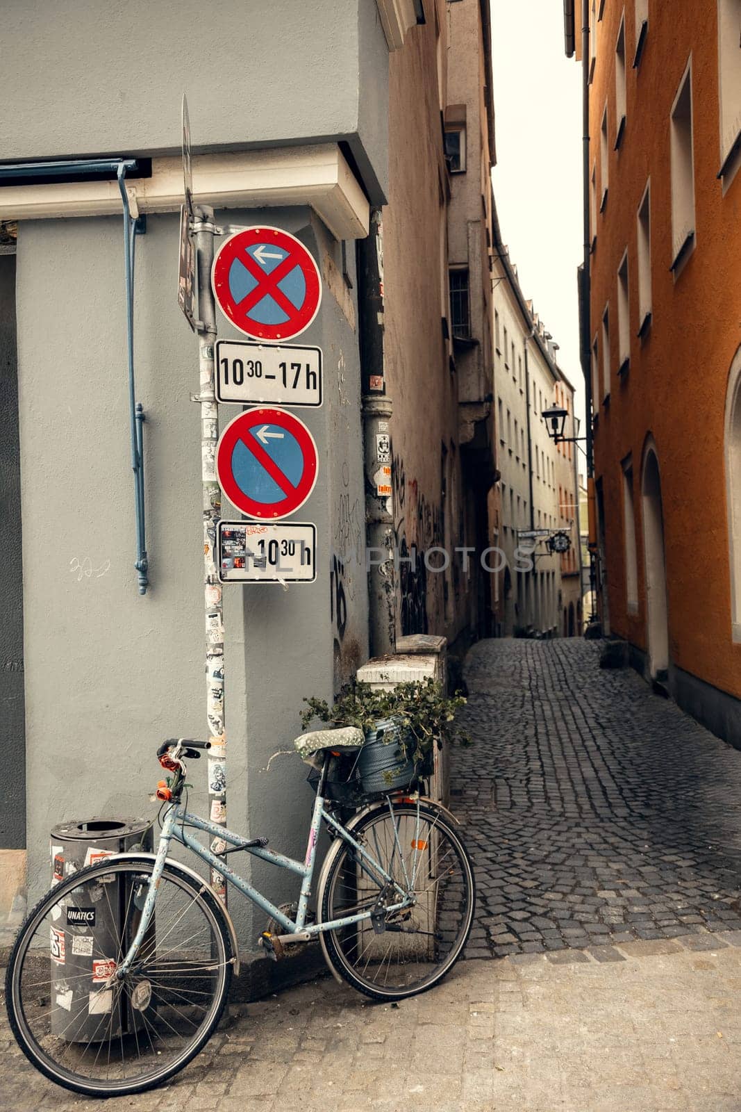 An old bicycle with a flower pot leans against a pole with traffic signs in a picturesque European alley. The cobblestone street and historic buildings create a nostalgic urban atmosphere