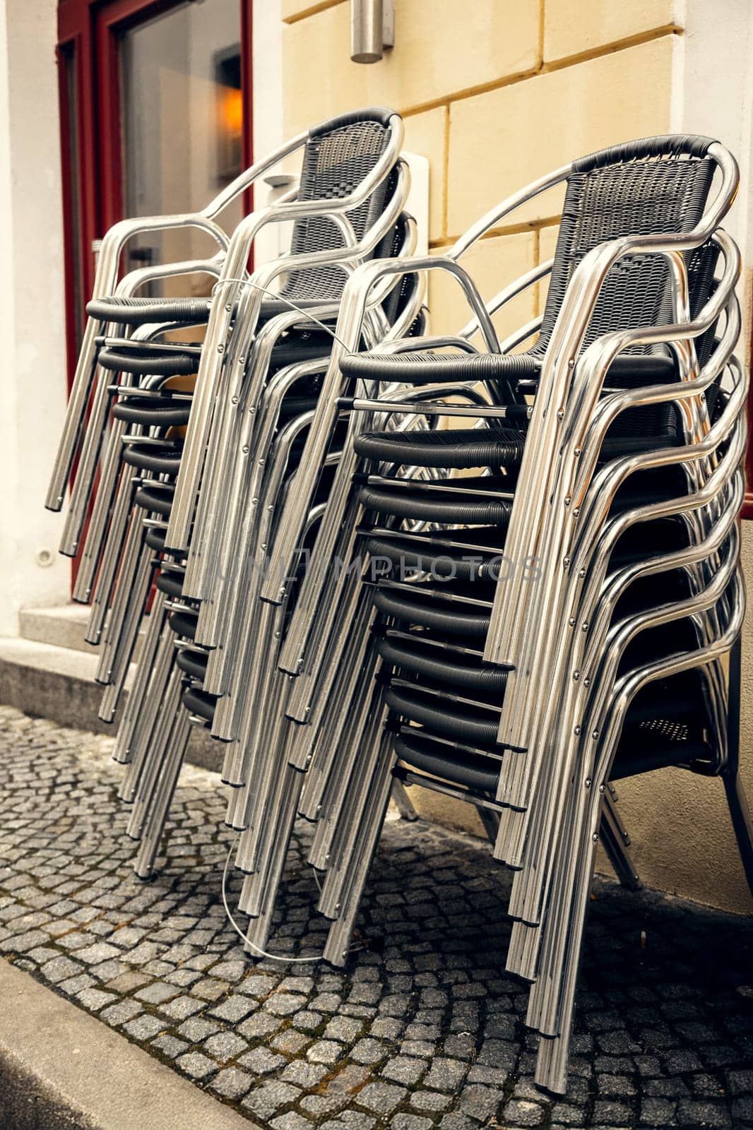 Stacked metal chairs outside European cafe. Neatly stacked metal and wicker chairs outside cafe, creating pattern of curved lines and reflections. Symbol of dining European street life