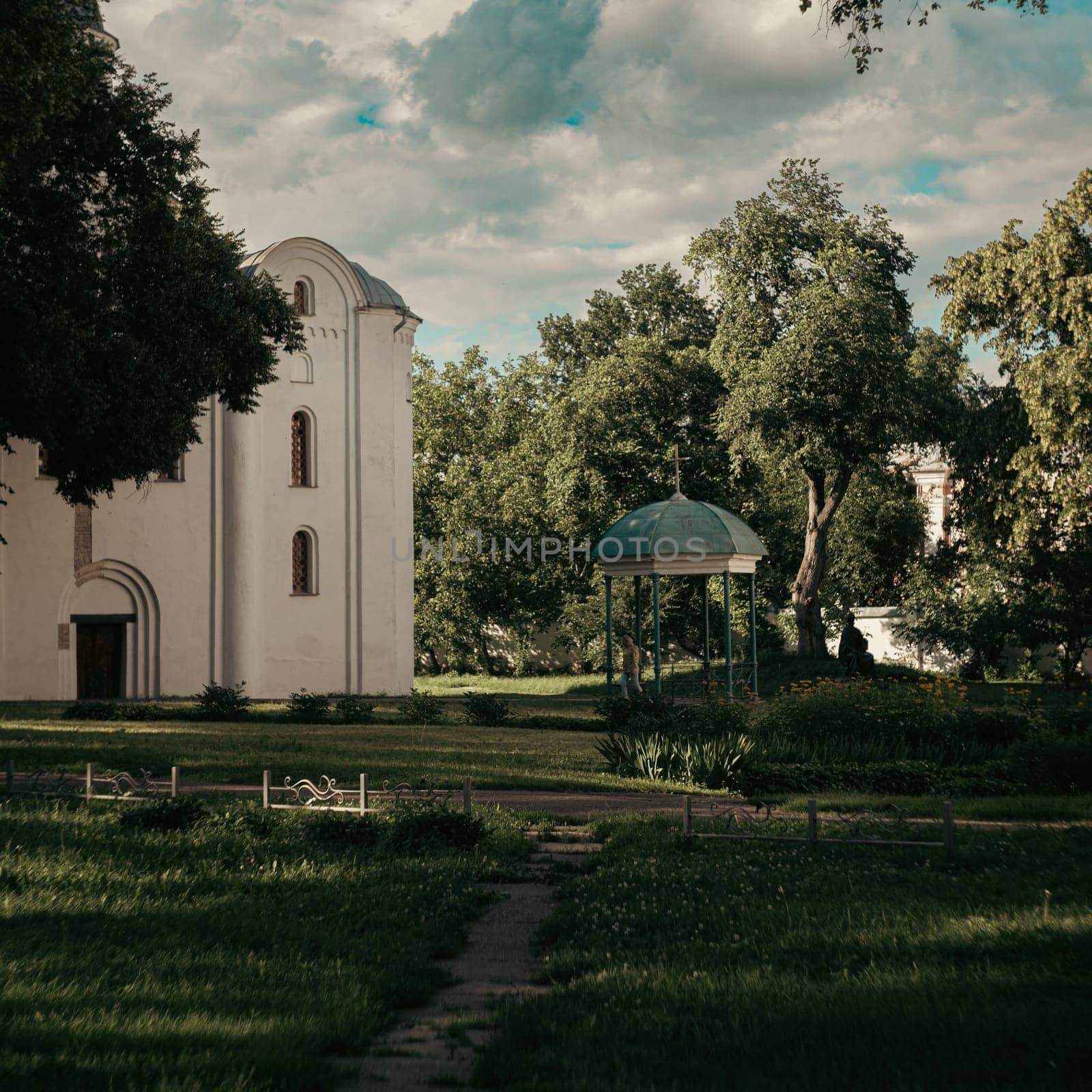 This scene shows a white church building with arched windows in a well-kept garden. A small gazebo is visible in the center of the garden, shaded by tall trees.