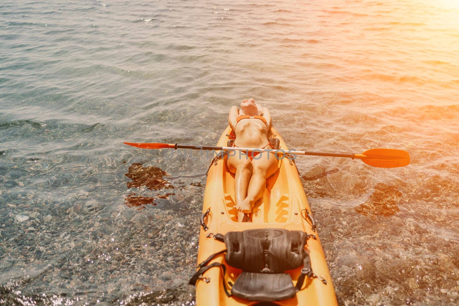 A woman is laying on a kayak in the water. The kayak is orange and has a paddle on it. The woman is wearing a red hat