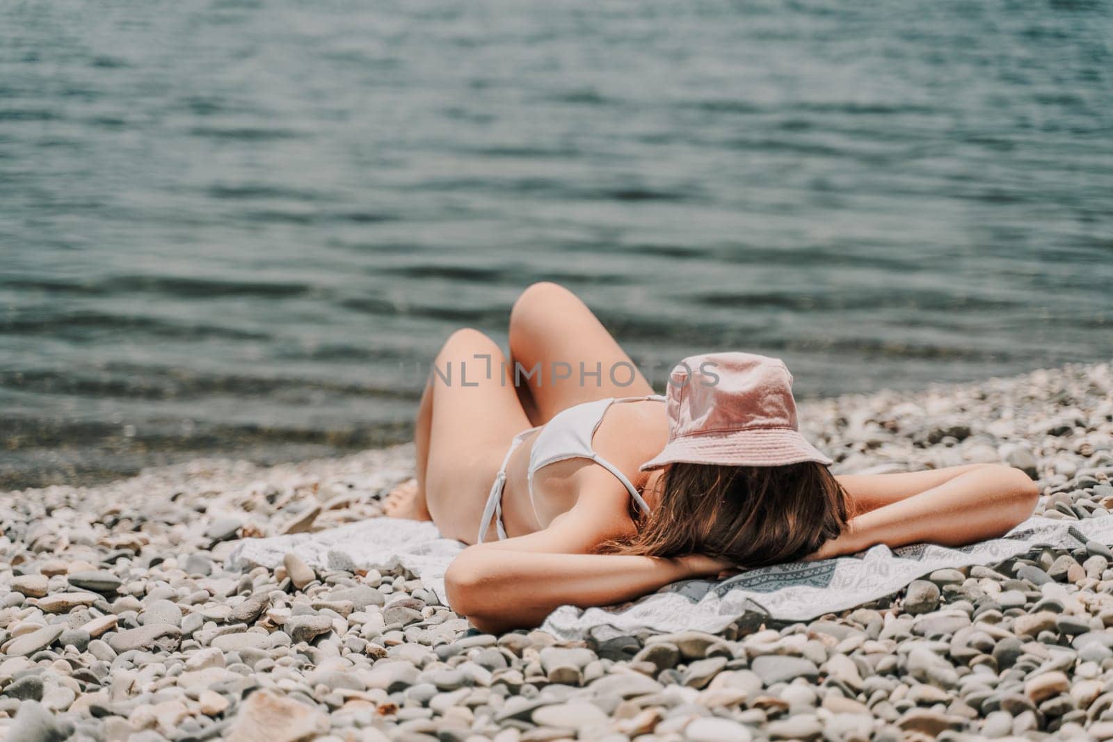 A woman is laying on the beach with a pink hat on her head. She is wearing a white bikini and is laying on a towel. The beach is rocky and the water is calm. Scene is relaxed and peaceful