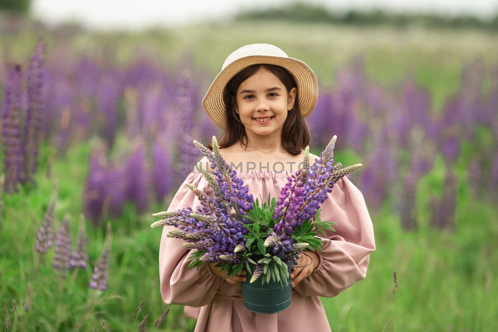 Young caucasian female child holding lupine flowers in field, wearing straw hat.