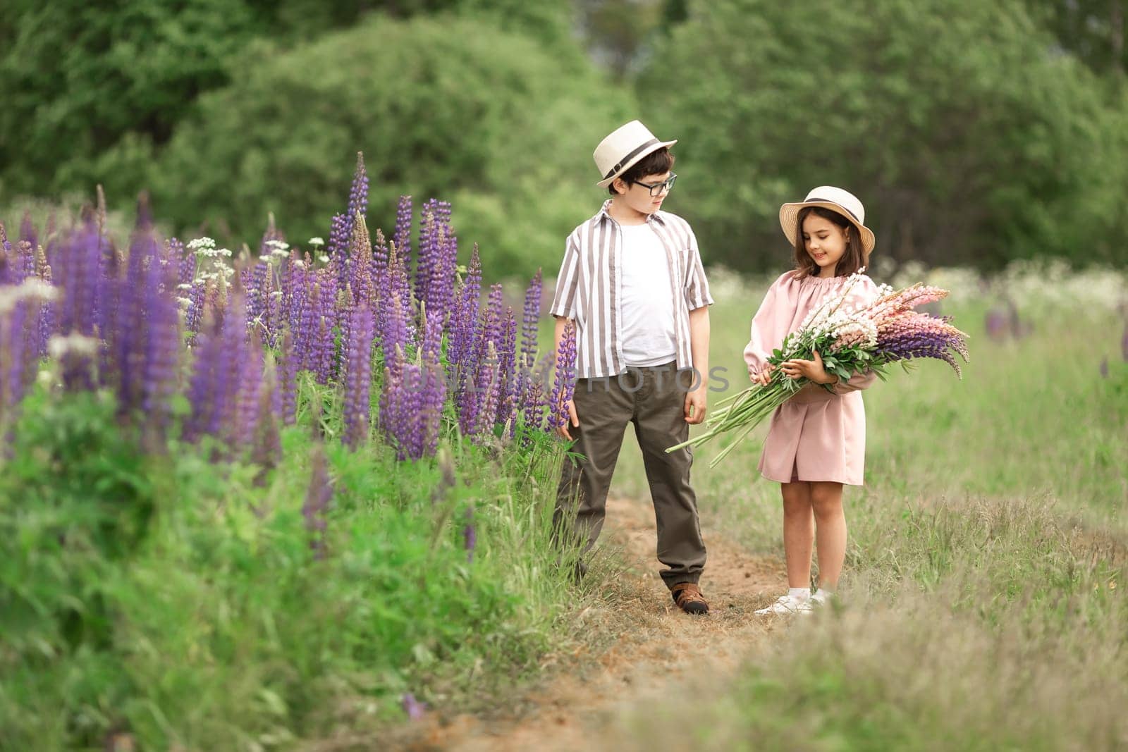Caucasian children in flower field: boy and girl with hats and bouquet of lupines.