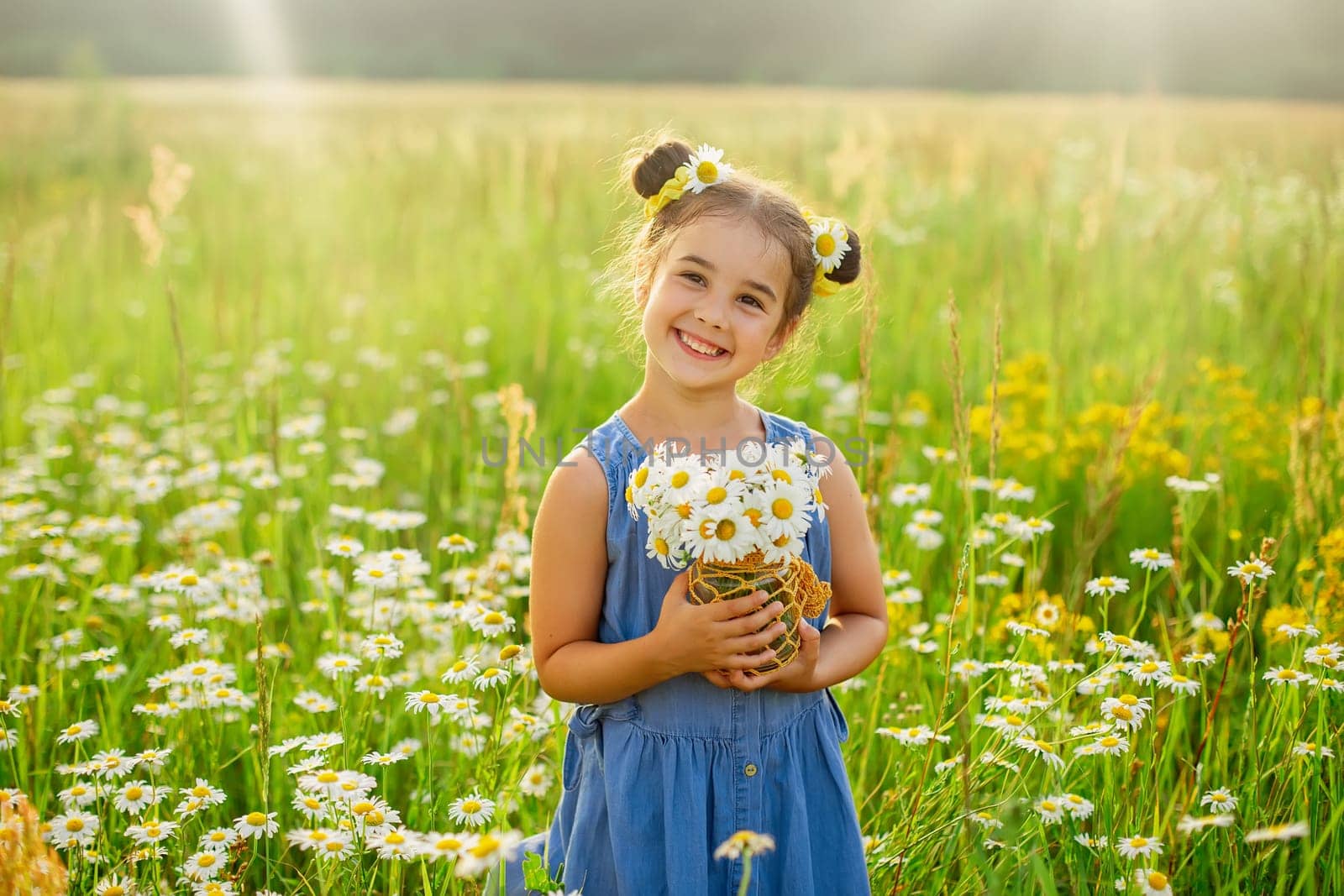 Cute smiling little girl, in a blue dress, holds a bouquet of daisies, stands in a field with daisies on a sunny day. Copy space
