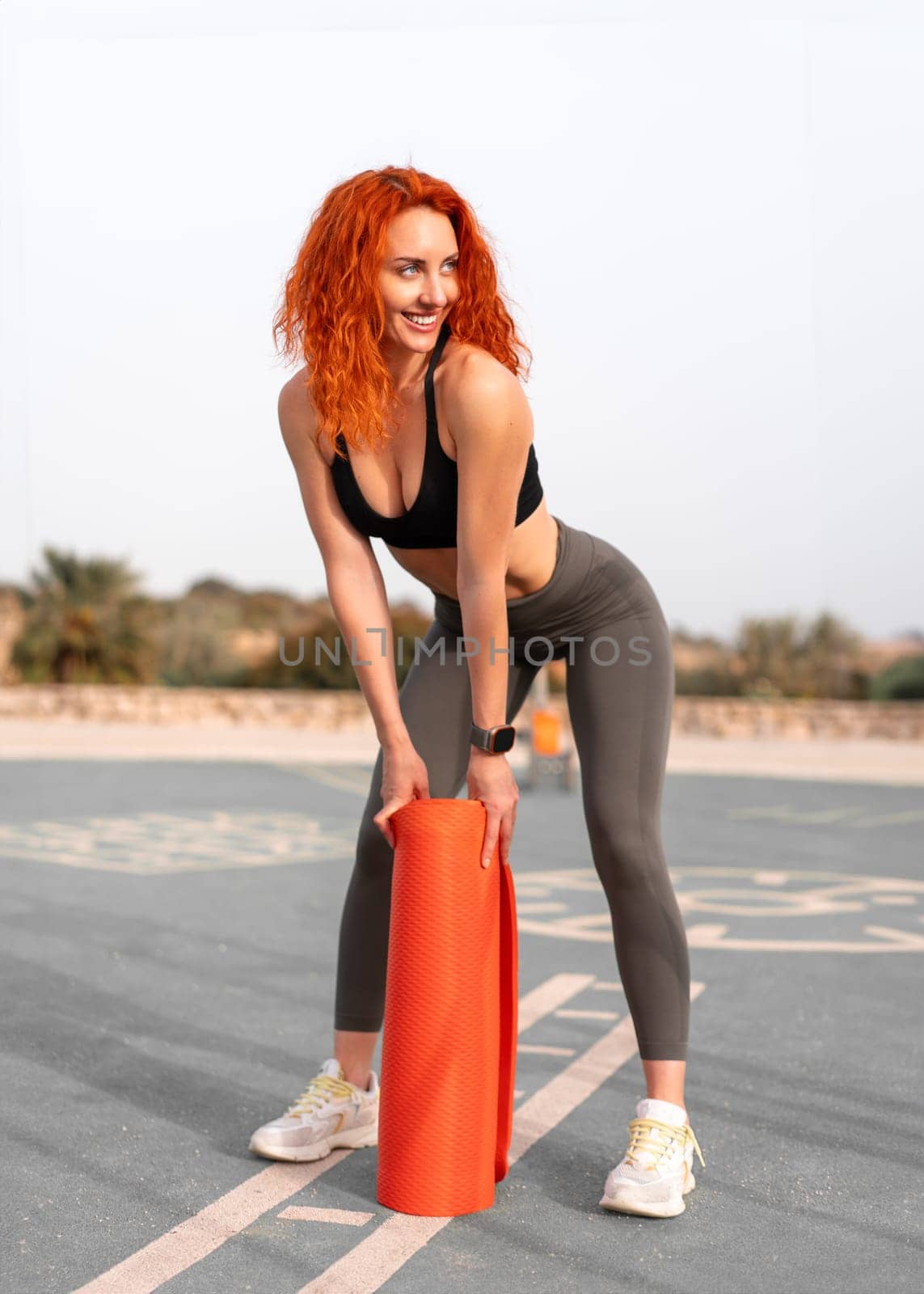 Sensuous jogger woman holding rolled up exercise mat at outdoor gym. Full body of fit young female in activewear is looking away while bending during daytime