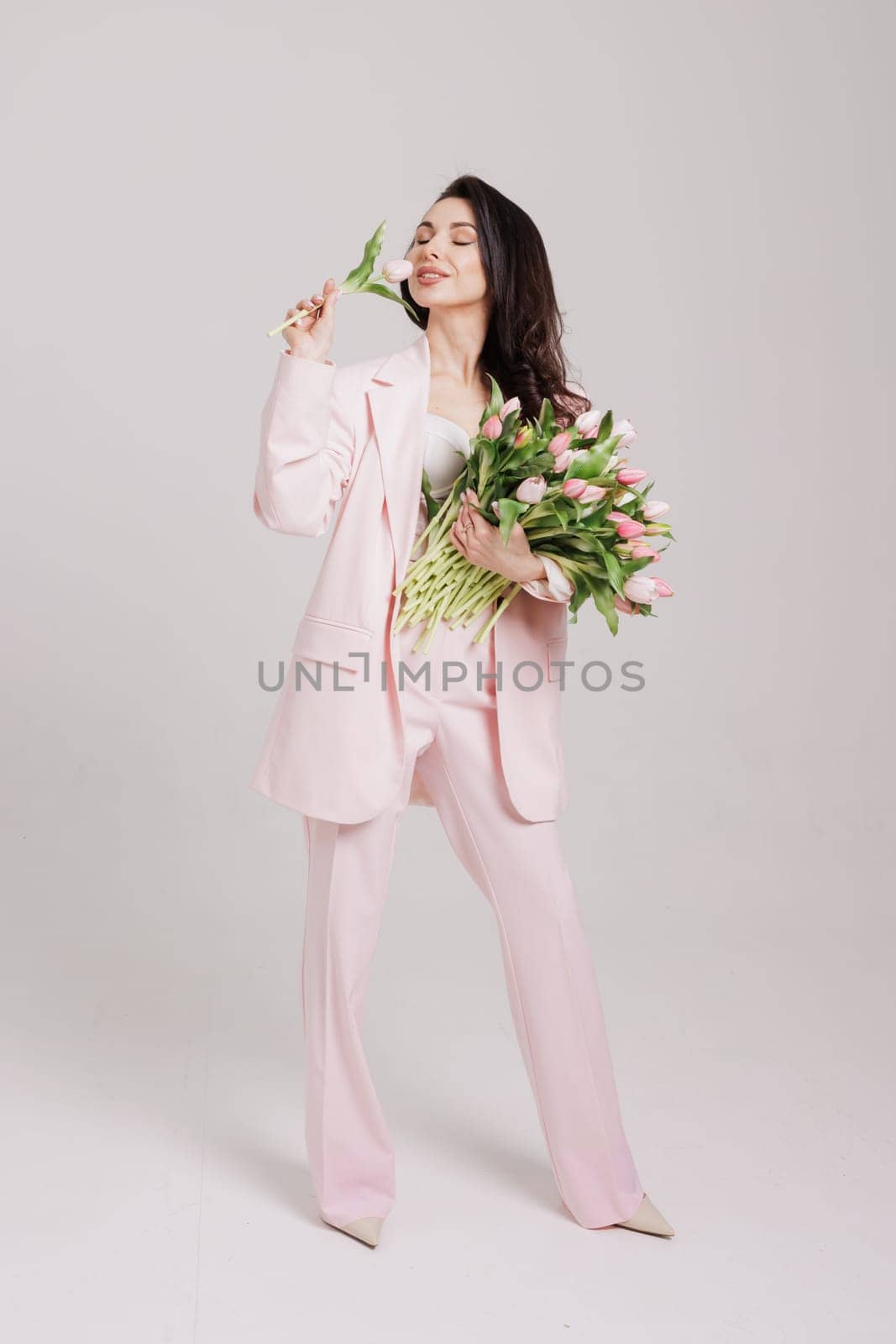 Smiling woman in a pink suit holding a bouquet of fresh tulips, posing against a neutral background in a soft, feminine, and elegant spring-inspired look