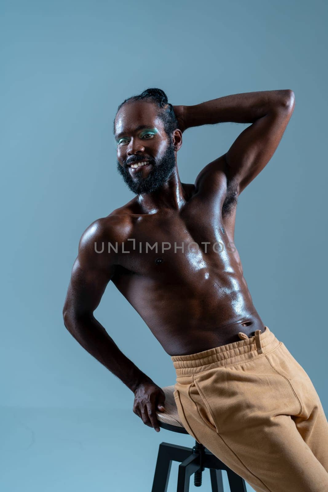 Happy shirtless black gay model leans casually on stool against blue background, showcasing confidence and joy with warm smile, exuding sense of positivity and self-expression