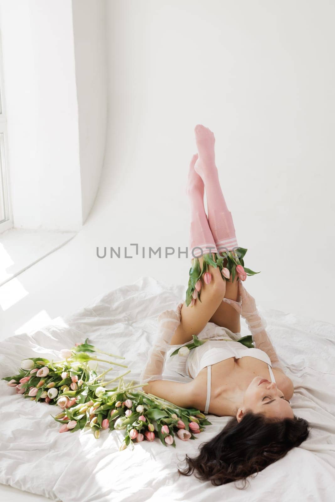 Elegant woman in lingerie lying on a bed with fresh tulips, wearing sheer gloves and pink socks, in a bright, minimalistic interior