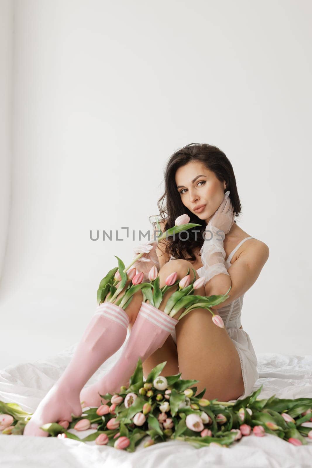 Sensual woman in white lingerie and pink socks sitting on bed with fresh tulips, in soft natural light.