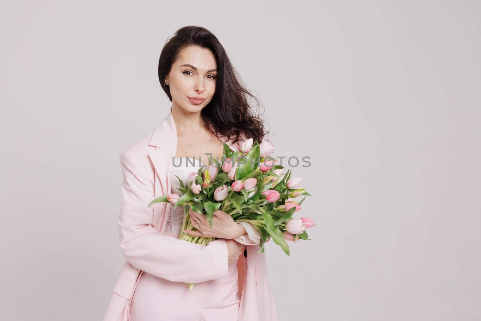 Smiling woman in a pink suit holding a bouquet of fresh tulips, posing against a neutral background in a soft, feminine, and elegant spring-inspired look