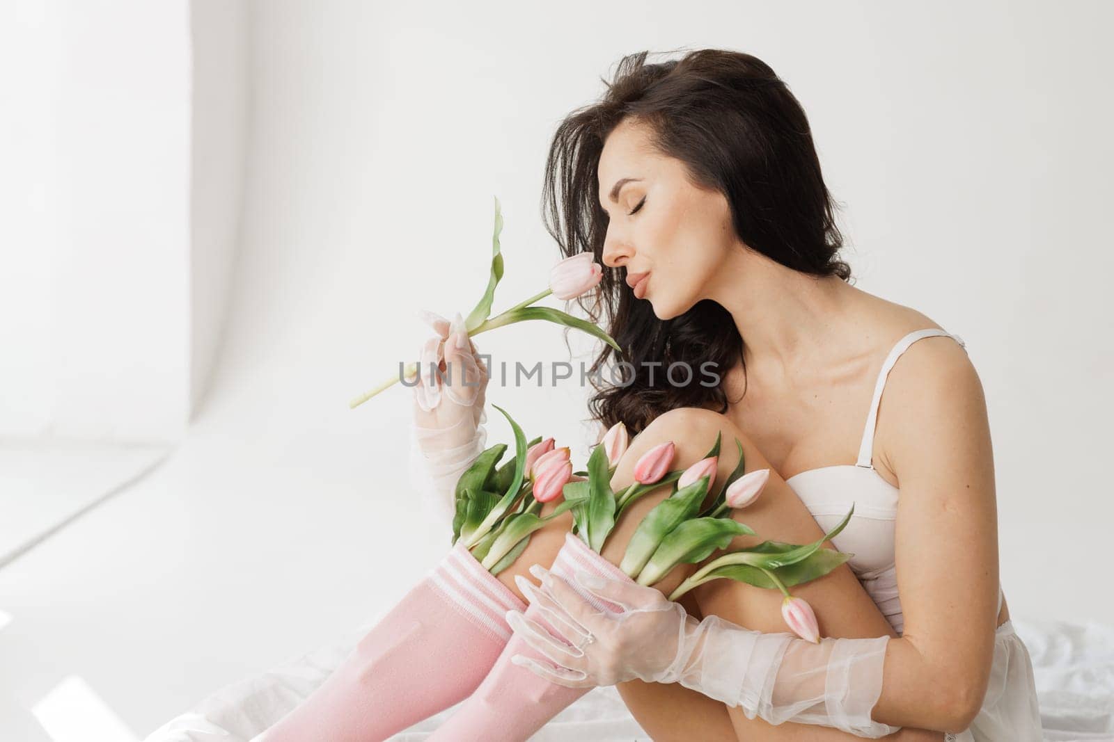 Sensual woman in white lingerie and pink socks sitting on bed with fresh tulips, in soft natural light.