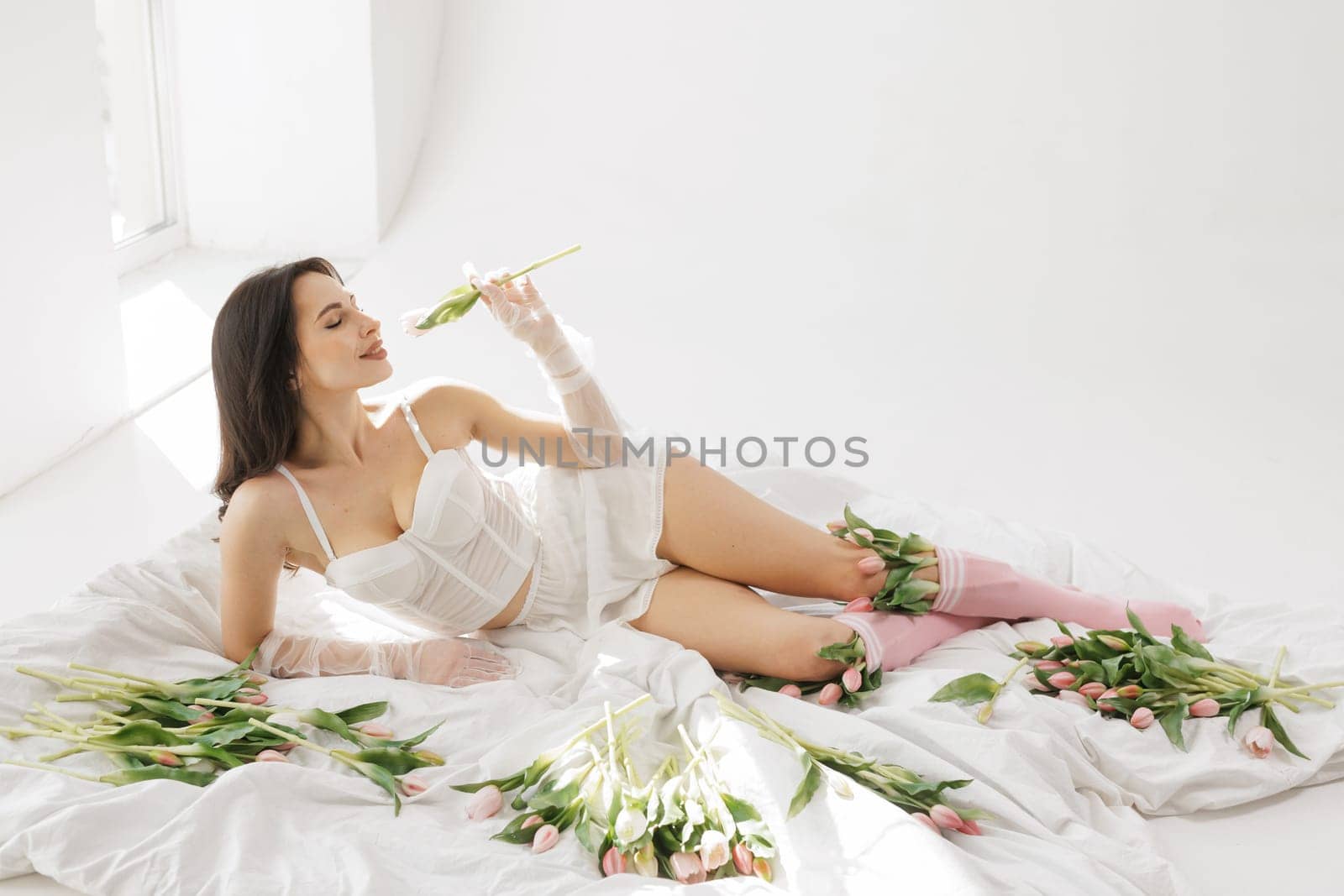 Elegant woman in white lingerie lying on bed with tulips, enjoying flowers in soft natural light, romantic and feminine aesthetic