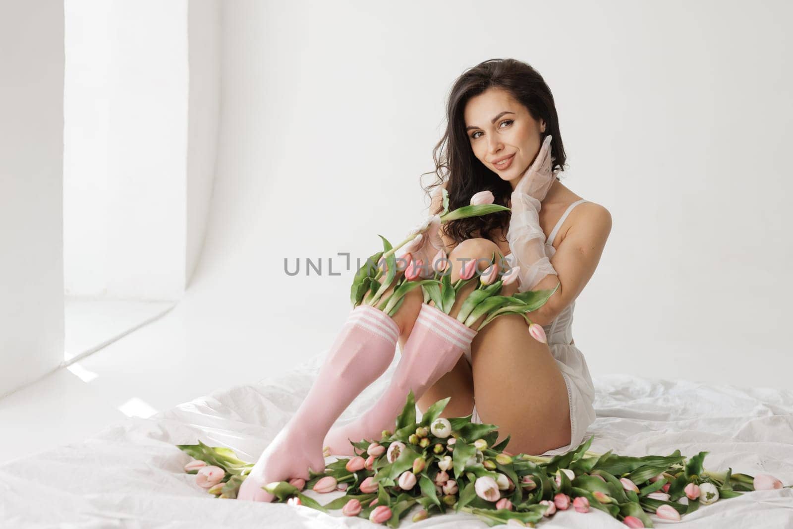 Sensual woman in white lingerie and pink socks sitting on bed with fresh tulips, in soft natural light.