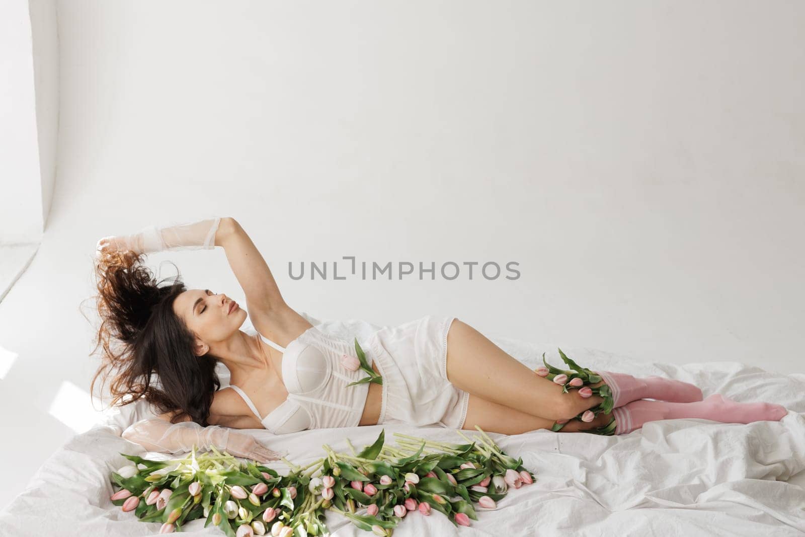 Elegant woman in lingerie lying on a bed with fresh tulips, wearing sheer gloves and pink socks, in a bright, minimalistic interior