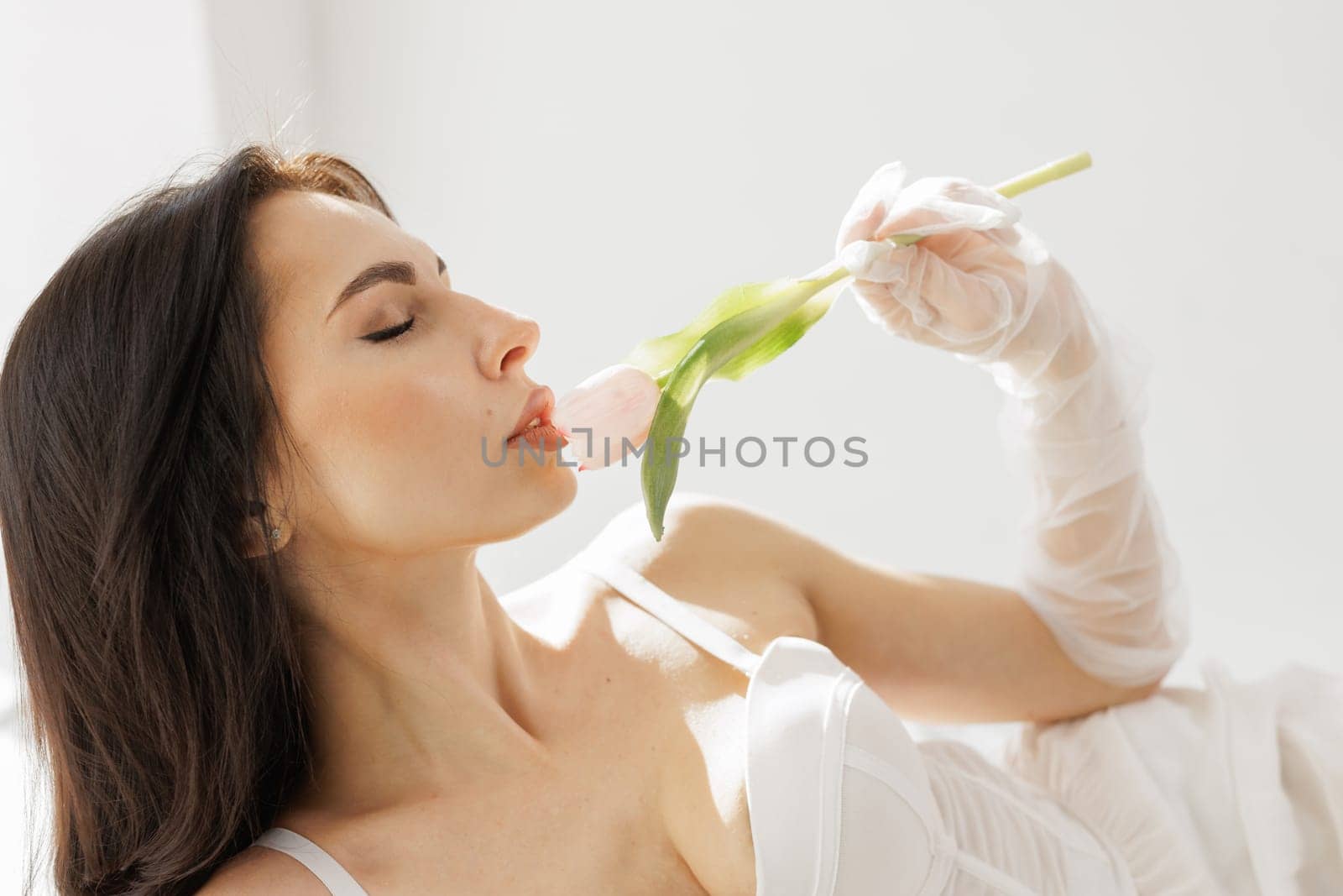 Elegant woman in white lingerie with delicate gloves holding a pink tulip near lips in soft natural light