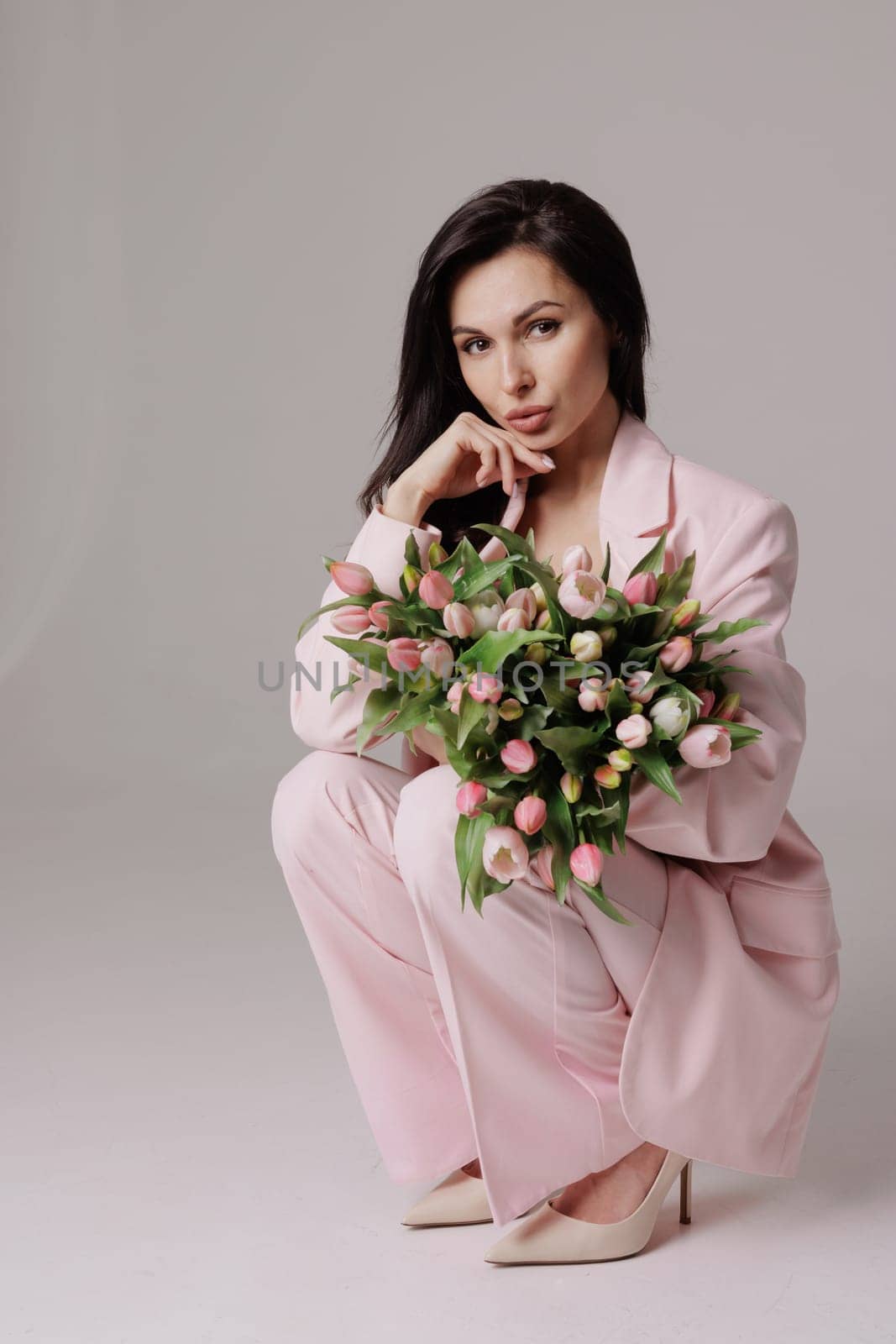 Smiling woman in a pink suit holding a bouquet of fresh tulips, posing against a neutral background in a soft, feminine, and elegant spring-inspired look