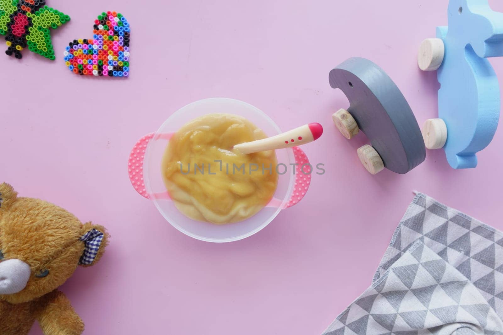 A bowl of baby food sits on a pink play mat surrounded by toys.