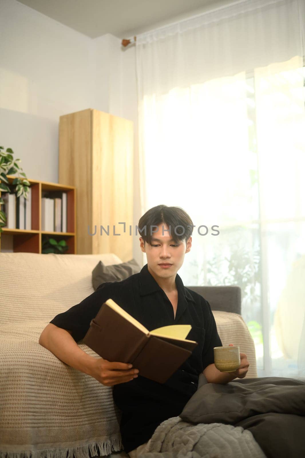 Young asian man reading a book while drinking hot drink in bright living room.