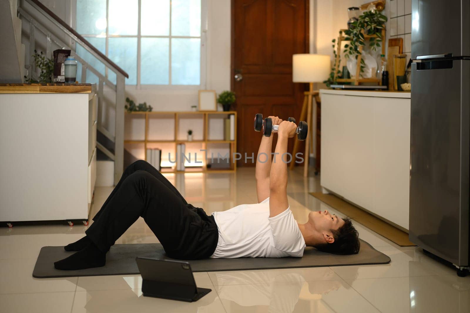 Young man lifting dumbbells on exercise mat and following an online workout session on digital tablet.