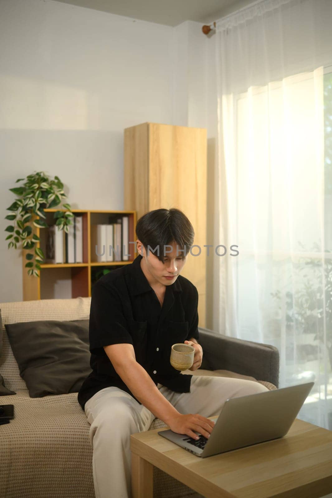 Young asian man holding a coffee cup and working on his laptop in a living room.