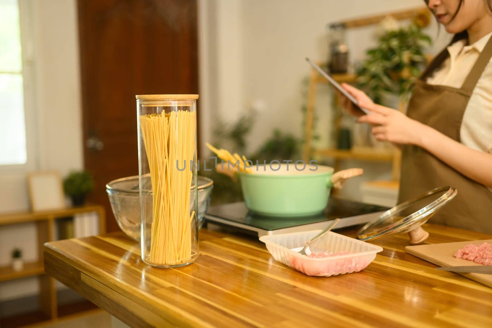 Dry spaghetti in a glass jar, meat, and boiling pot on kitchen counter with woman checking recipe on digital tablet.