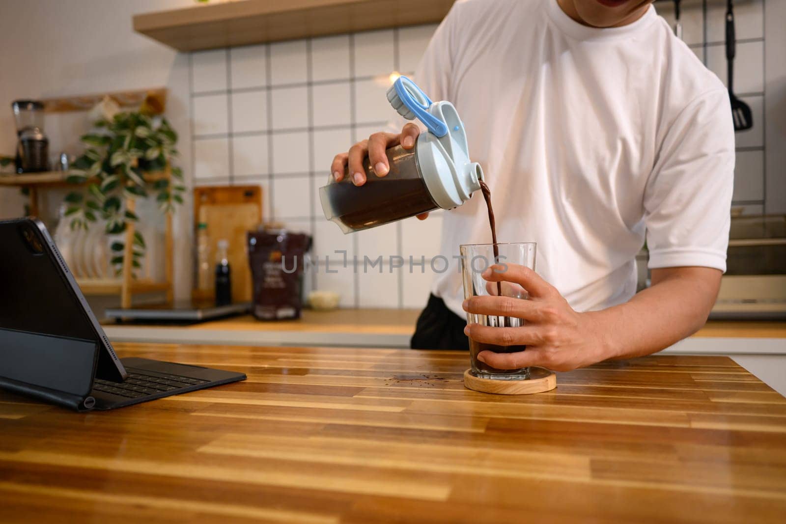 Man pouring mixed chocolate protein shake from a shaker bottle into a glass in a modern kitchen.