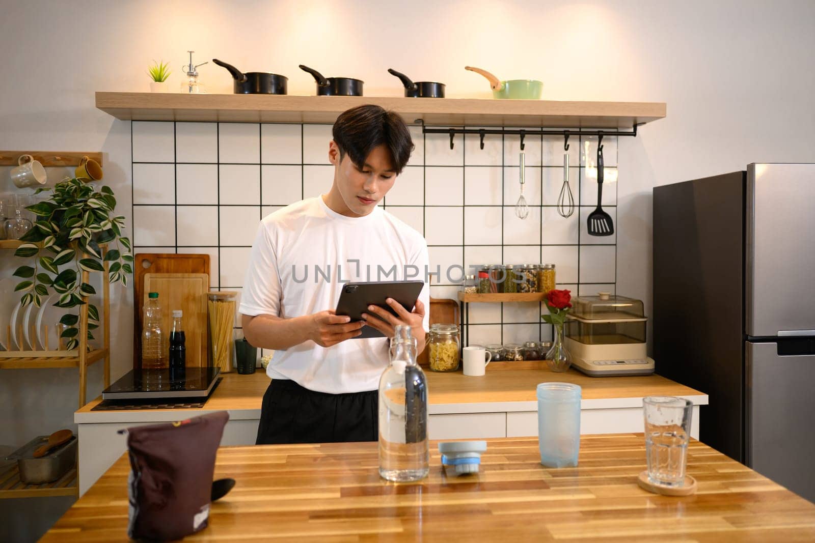 Young man wearing a white sports shirt, checking digital tablet while preparing a protein shake on kitchen counter.