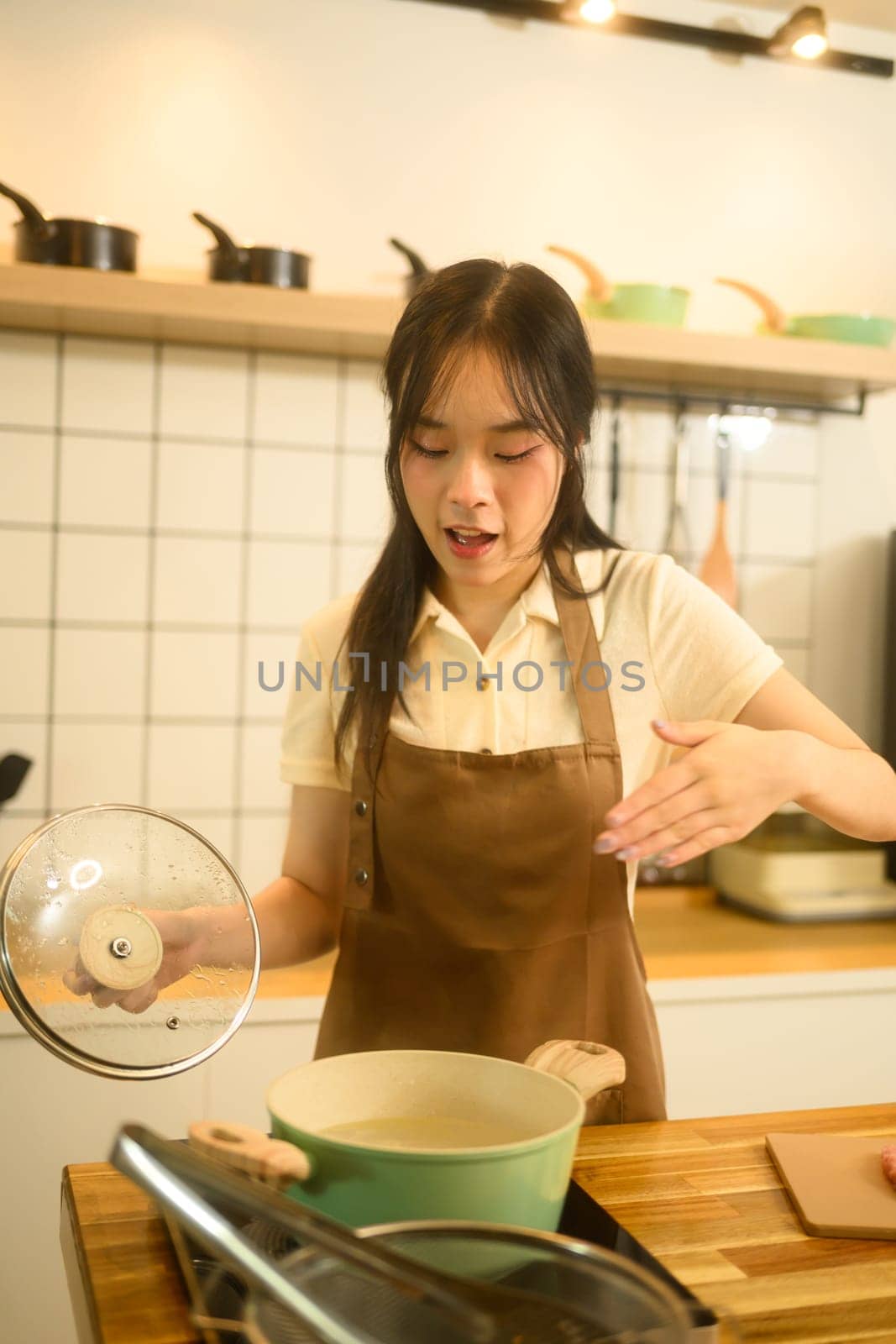 Young woman in brown apron lifting the lid off a pot while checking on boiling pasta.