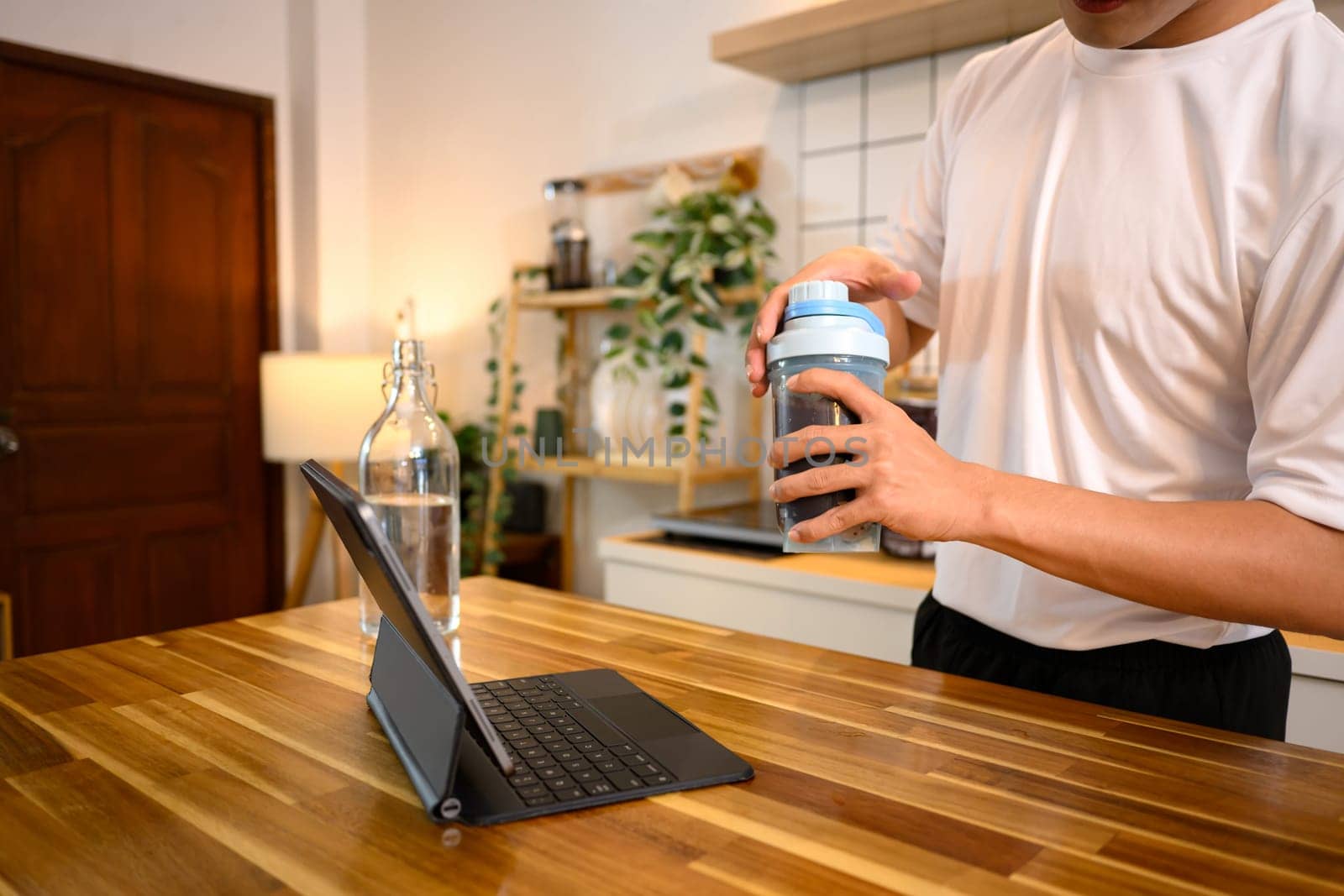 Young man wearing a white sports shirt, checking digital tablet while preparing a protein shake on kitchen counter.