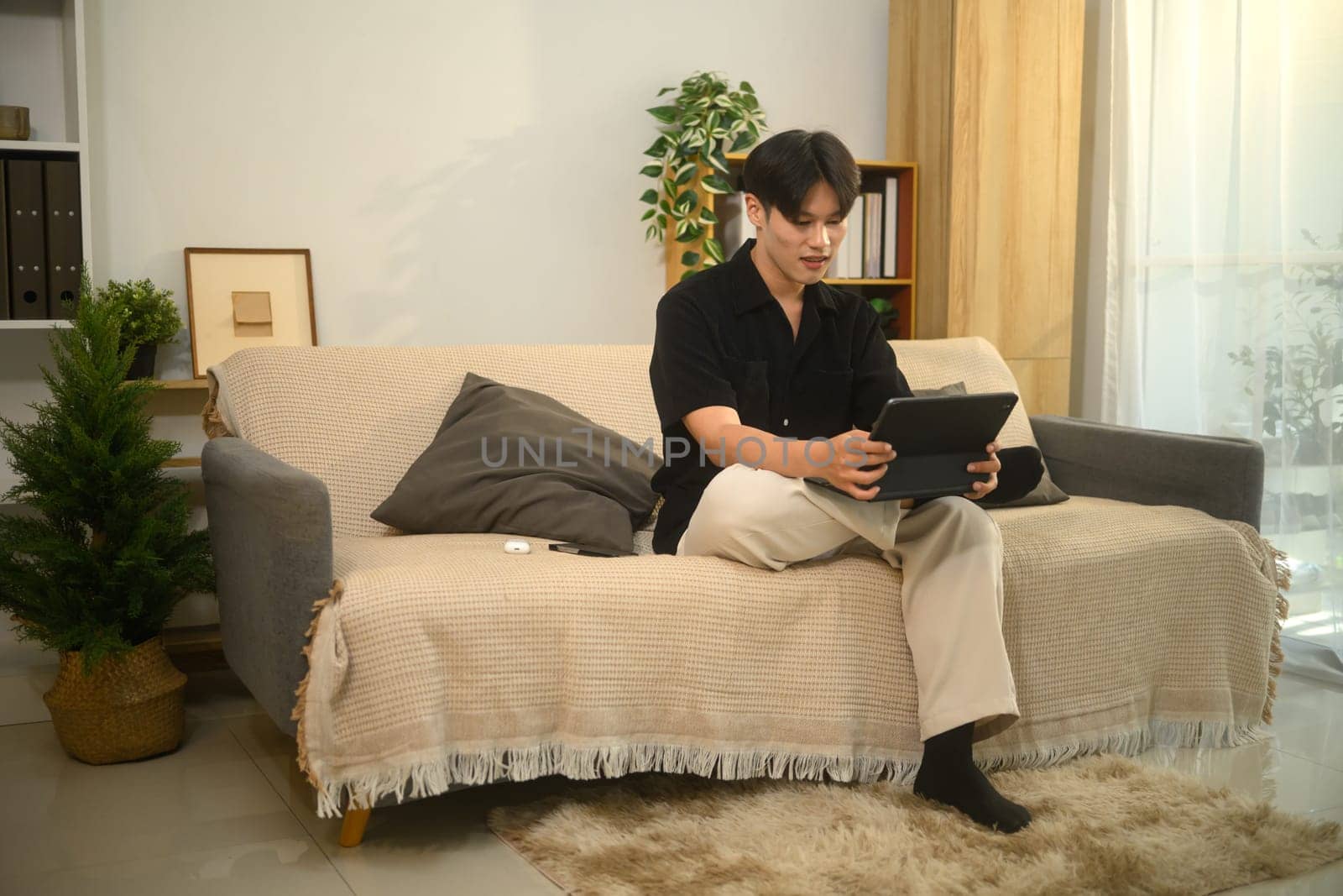Relaxed asian man sitting on cozy sofa in a modern living room and using digital tablet.