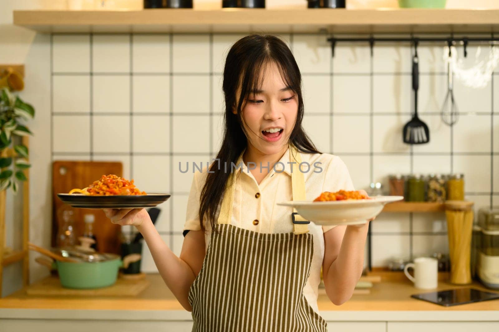 Cheerful woman holding two plates of homemade spaghetti standing in modern kitchen.