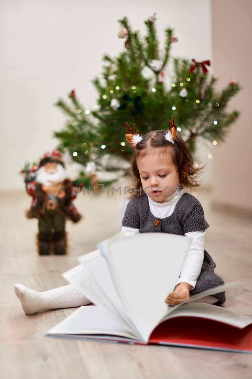 Cute little child girl reads a book near the Christmas tree at home