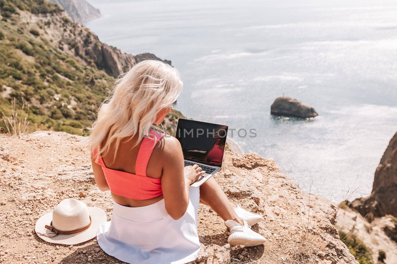 A woman is sitting on a rocky hillside with a laptop open in front of her. She is wearing a pink top and a straw hat. Concept of relaxation and leisure