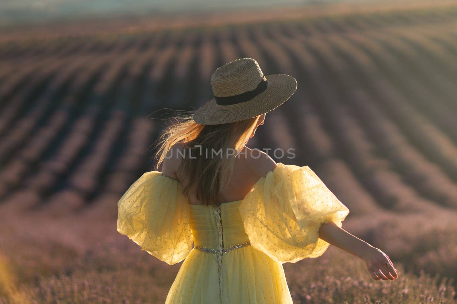 Here's a caption under 150 characters describing the image: Woman in yellow dress and straw hat, back to camera, stands in a vast lavender field at sunset.