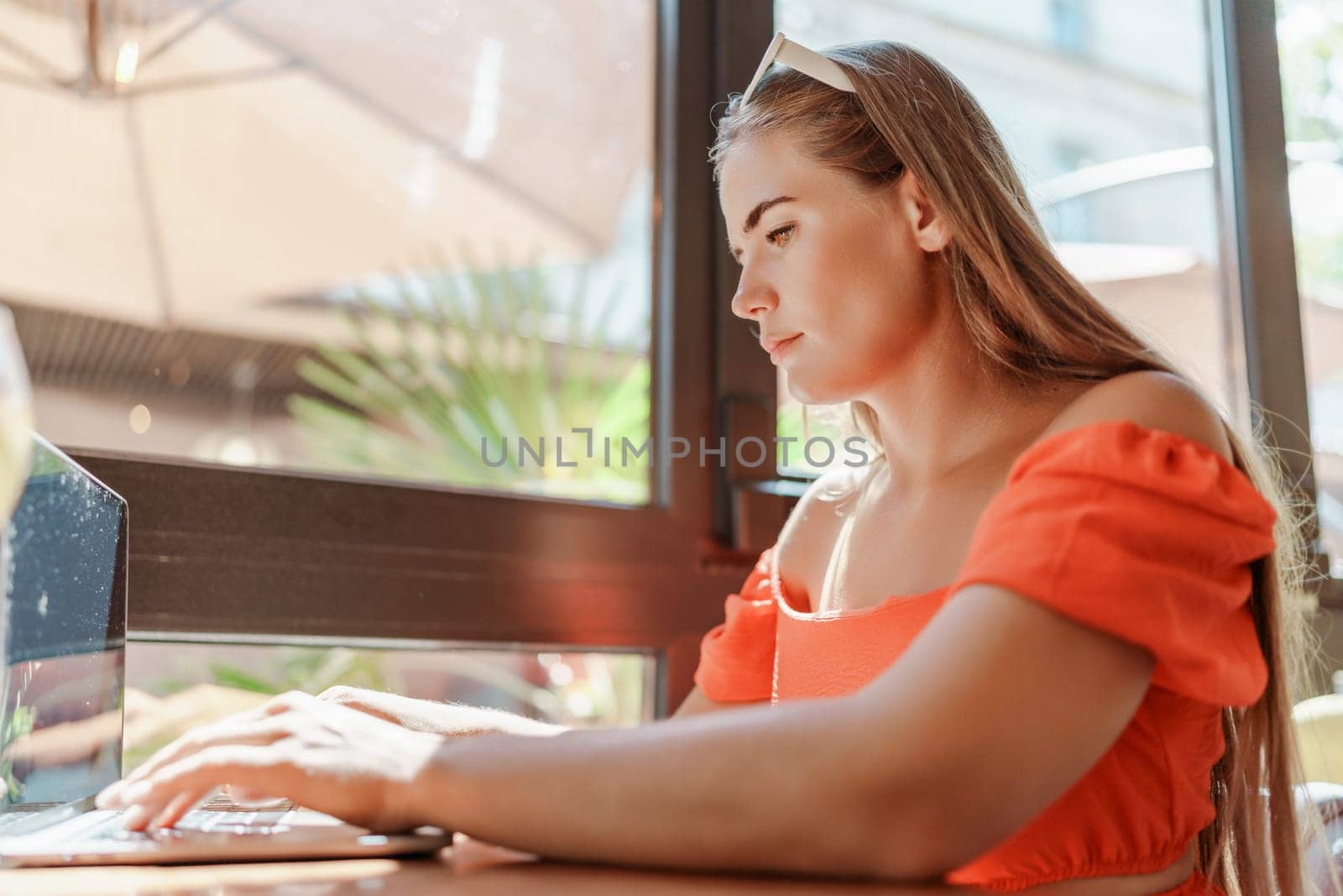 Woman works on laptop in a sunny cafe, glass of juice nearby. The setting is modern, with natural light illuminating the space. The image captures a moment of focused work