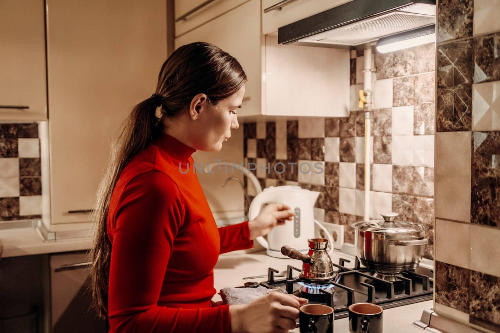 Woman in red turtleneck prepares coffee using a stovetop cezve and electric kettle in a domestic kitchen.